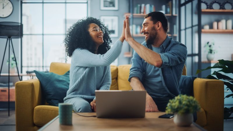 Diverse Multiethnic Couple are Sitting on a Couch Sofa in Stylish Living Room and Choosing Items to Buy Online with Laptop Computer, Give High Five. Friends or Colleagues are Discuss Work Projects.