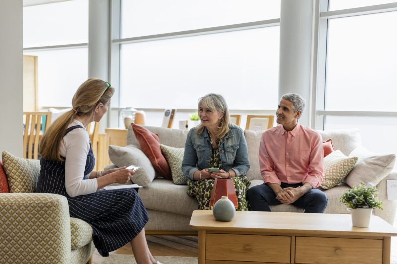 A mature couple sitting on a sofa in a furniture store, talking to a store employee about buying new furniture for their home. The employee is using a digital tablet while they talk.