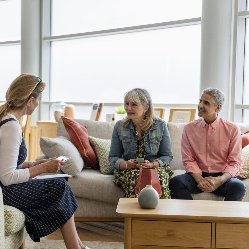 A mature couple sitting on a sofa in a furniture store, talking to a store employee about buying new furniture for their home. The employee is using a digital tablet while they talk.