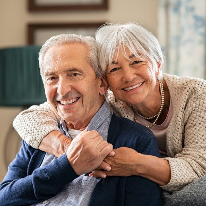 Portrait of romantic senior man with his beautiful wife stay at home. Smiling and caring old woman embracing from behind her retired husband sitting on couch. Cheerful old couple looking at camera with joy.