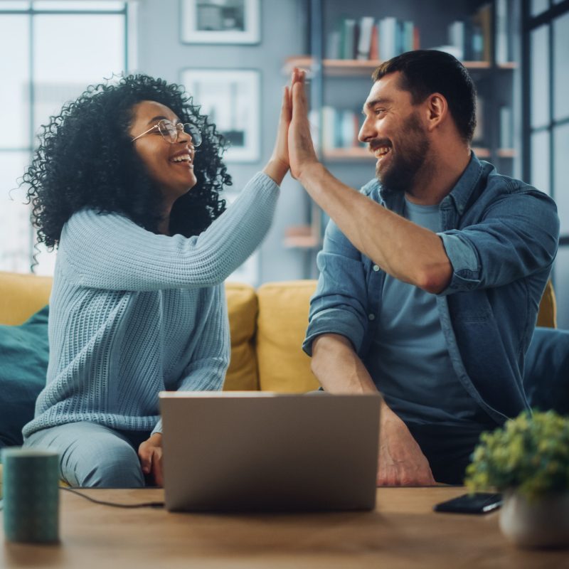 Diverse Multiethnic Couple are Sitting on a Couch Sofa in Stylish Living Room and Choosing Items to Buy Online with Laptop Computer, Give High Five. Friends or Colleagues are Discuss Work Projects.