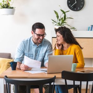 Happy husband and wife read good news online at laptop, smiling man holding documents receiving positive decision from bank