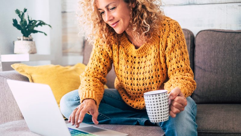 Nice beautiful lady with blonde curly hair work at the notebook sit down on the sofa at home - check on line shops for cyber monday sales - technology woman concept for alternative office freelance